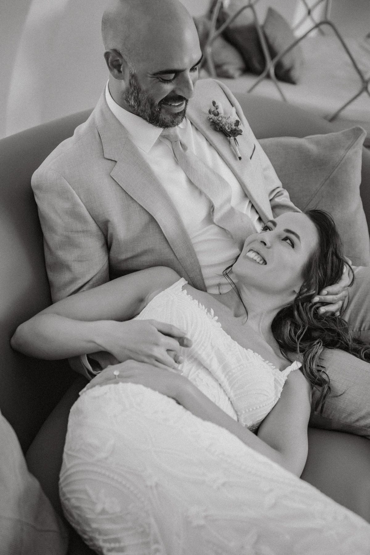 Black and white photo of a bride and groom sharing a tender moment on a sofa, smiling at each other.