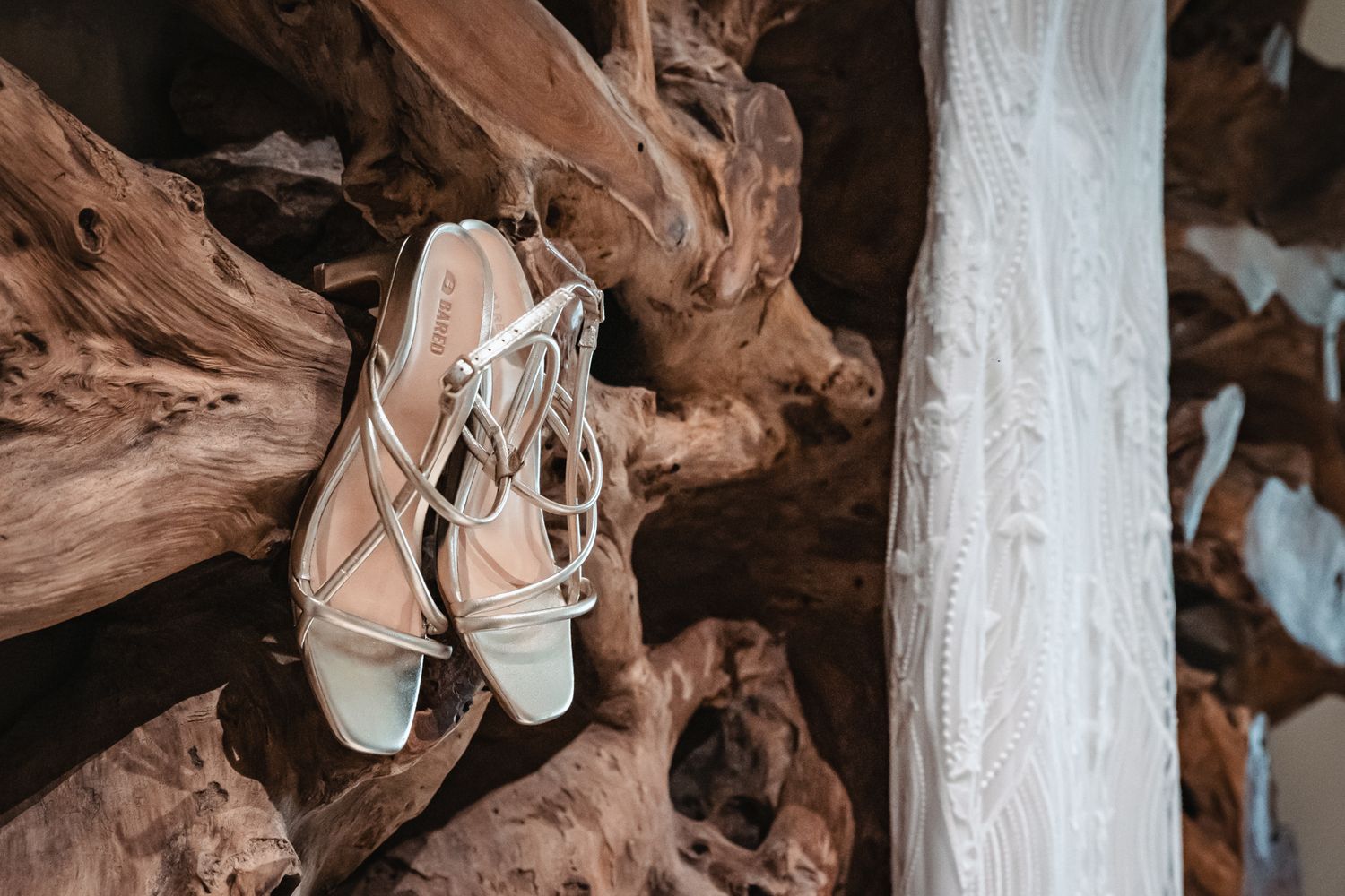 Elegant white strappy bridal heels hanging on rustic driftwood beside a lace wedding dress.