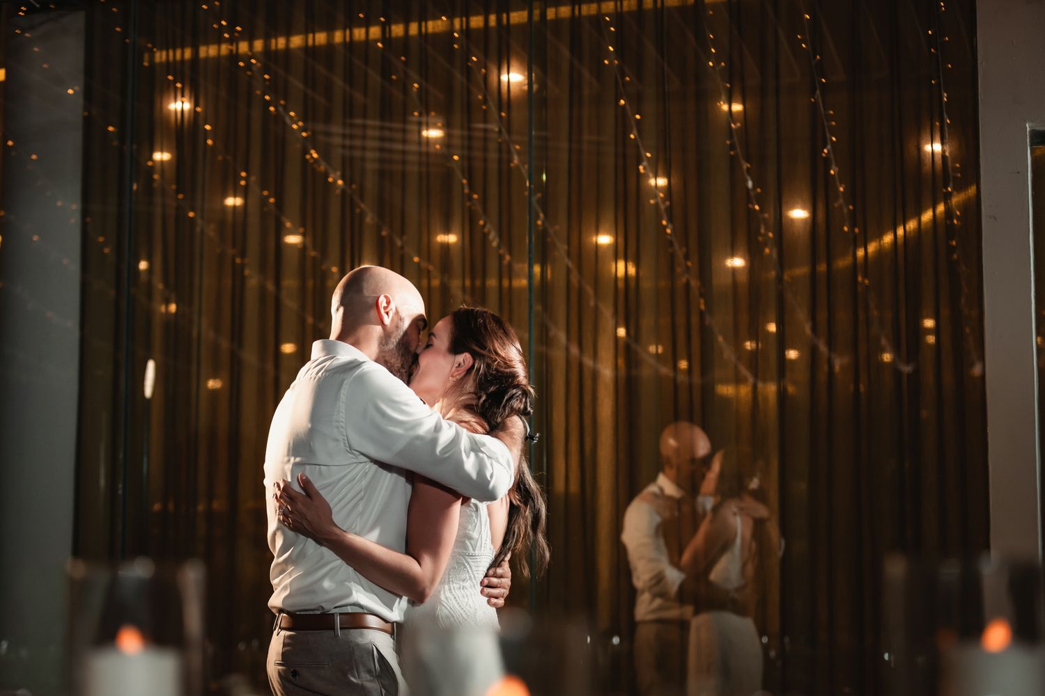Couple shares first dance kiss under warm string lights at romantic wedding reception.