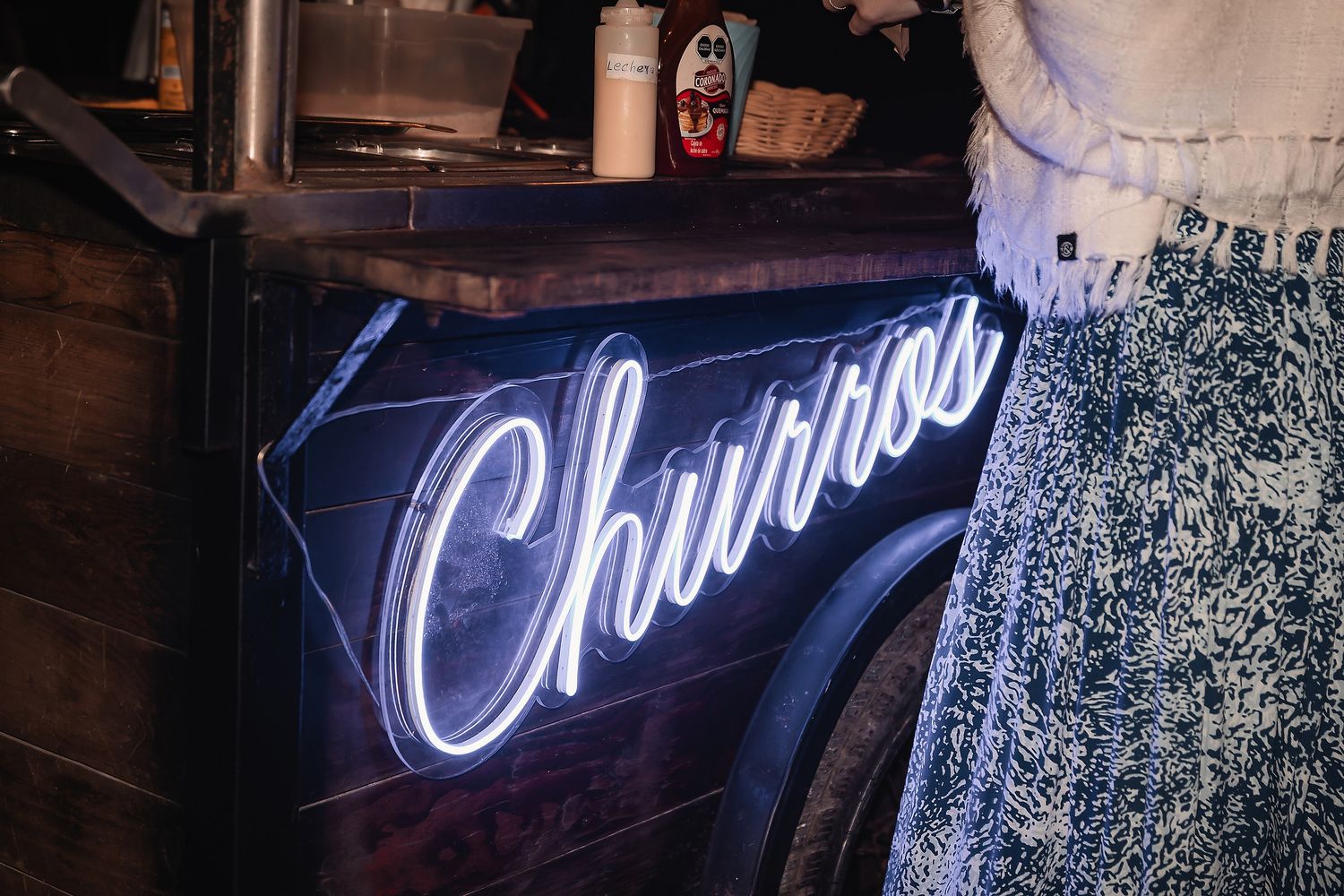 Glowing neon Churros sign on a dark wooden cart at a food stall, with a person in a floral skirt standing nearby.