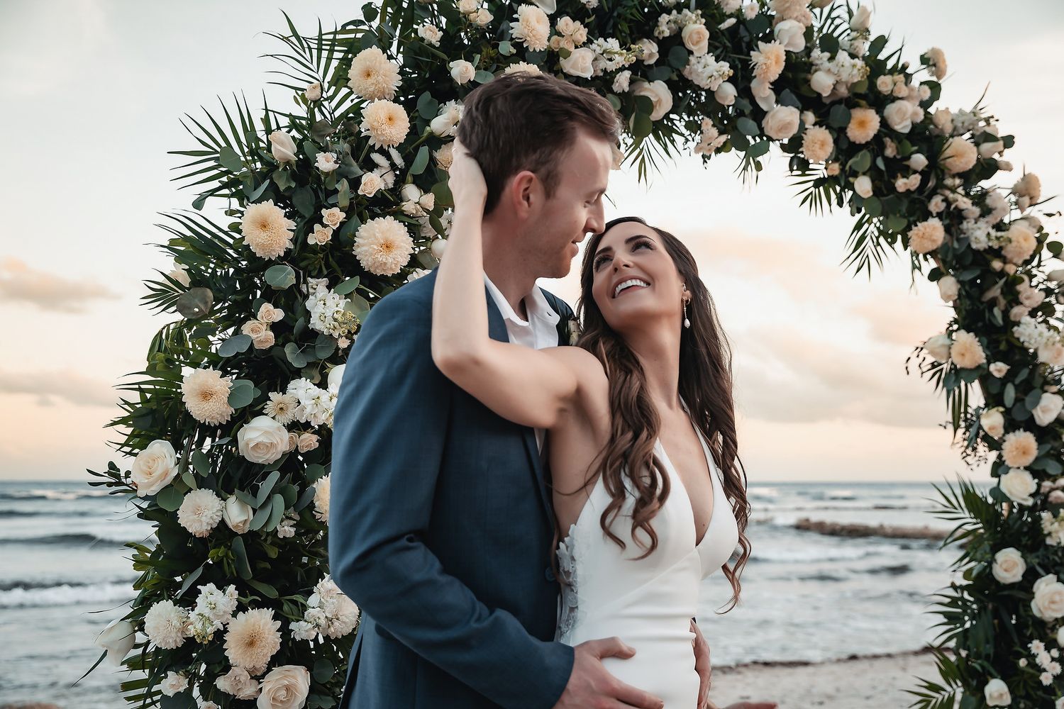Bride and groom embracing under a white floral arch on a beach at sunset.