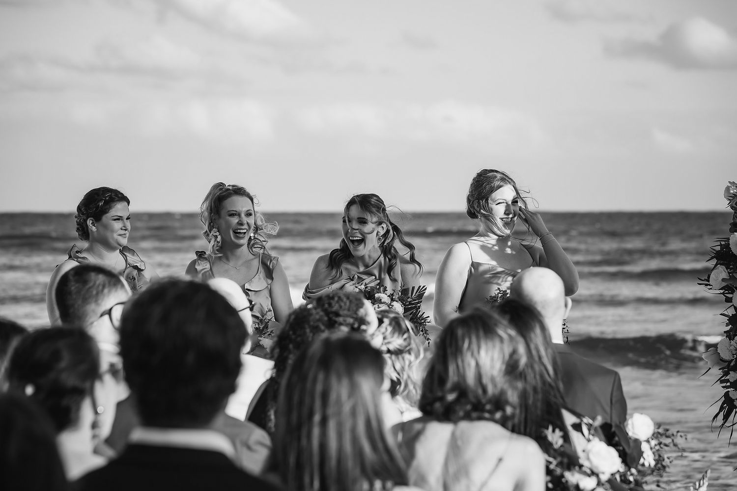Black and white beach wedding ceremony with bridesmaids holding bouquets, ocean waves in the background.