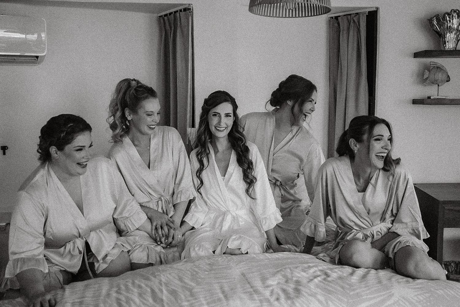 Bridesmaids in silk robes laughing and sitting together on a bed during wedding morning preparations.