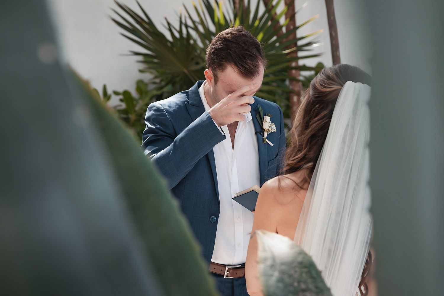 Emotional groom wiping tears during outdoor wedding ceremony surrounded by tropical greenery.
