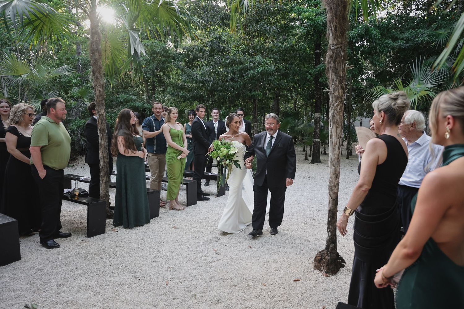 Bride and groom walk down sandy aisle surrounded by tropical palms with guests standing at outdoor ceremony.