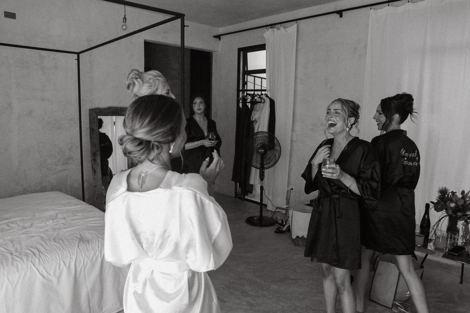 Black and white photo of bridesmaids in robes laughing and getting ready together in a bright bedroom.
