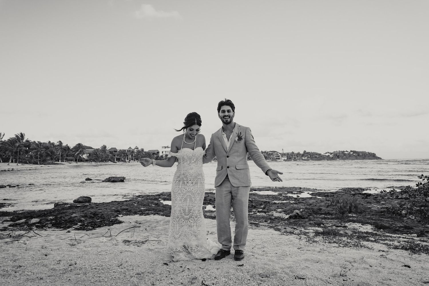 Joyful bride and groom pose on rocky beach in black and white wedding photo.