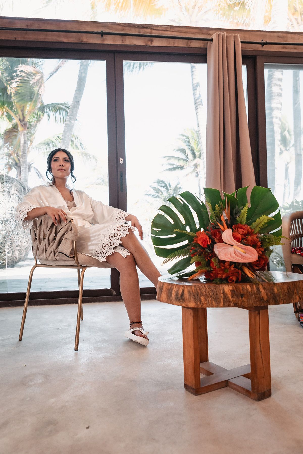 Bride in white lace robe sits elegantly in chair beside tropical floral arrangement with monstera leaves and anthuriums.