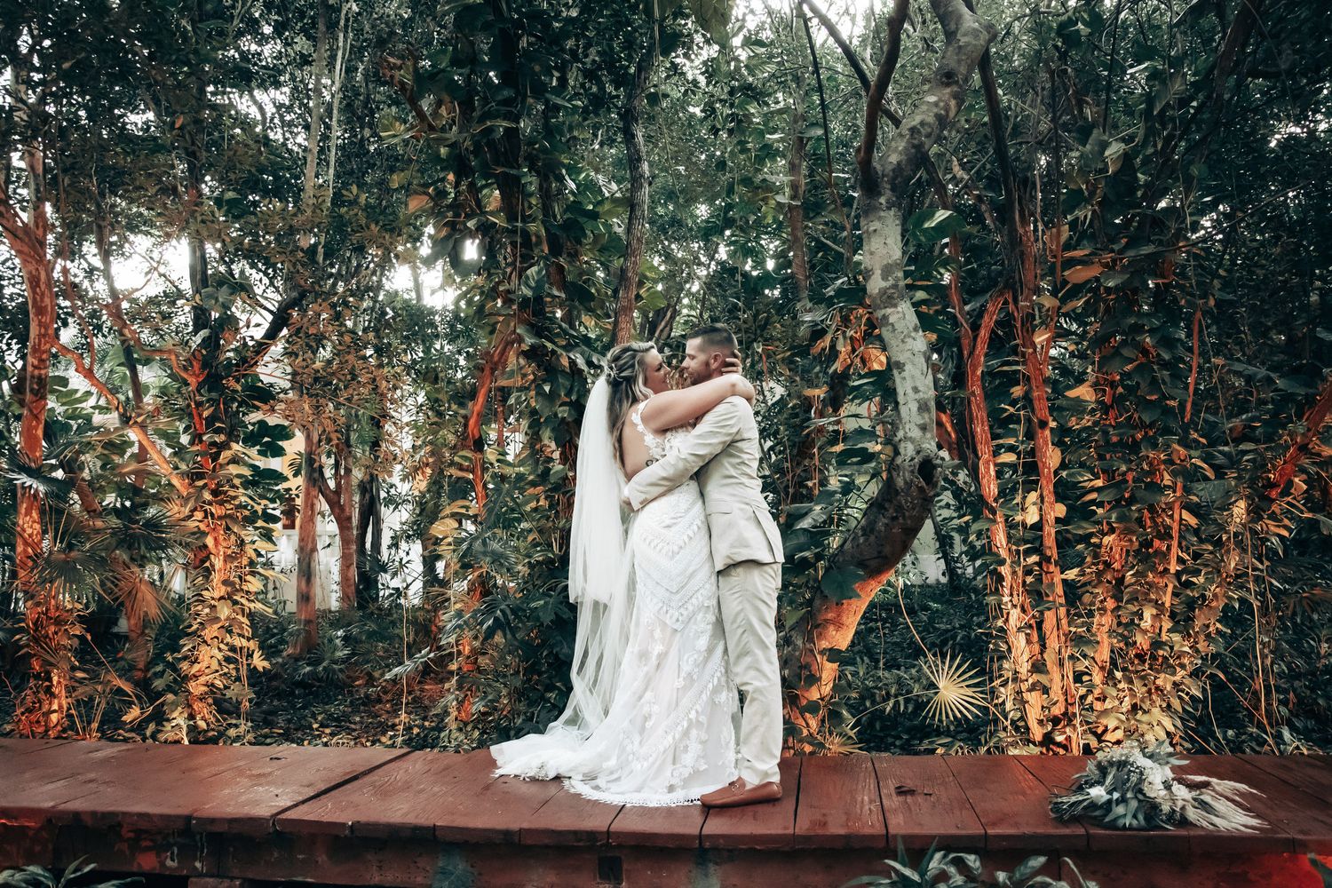 Bride and groom embracing on a wooden deck surrounded by lush tropical forest.