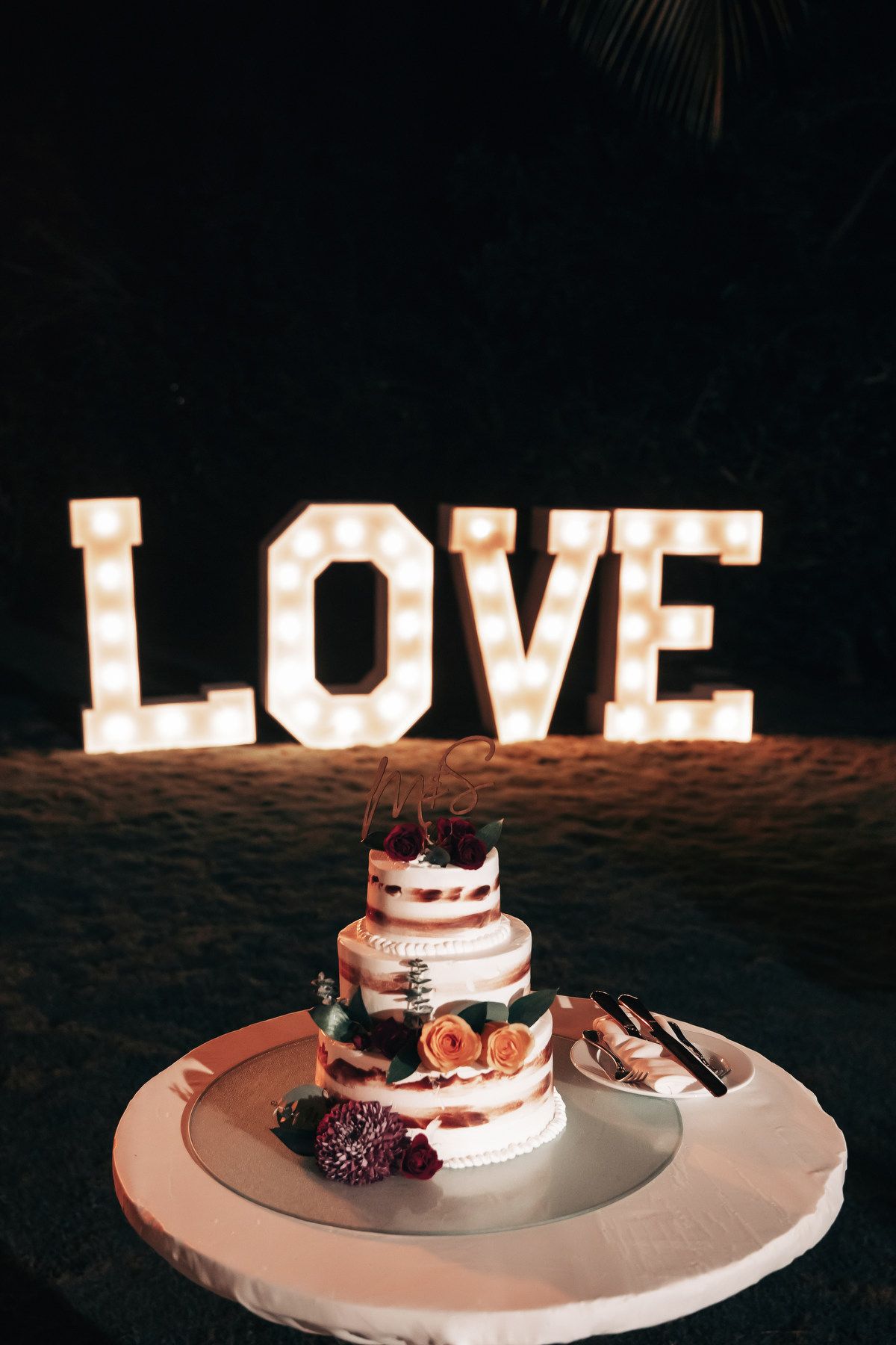 Two-tier wedding cake with floral decor displayed in front of illuminated LOVE marquee sign.