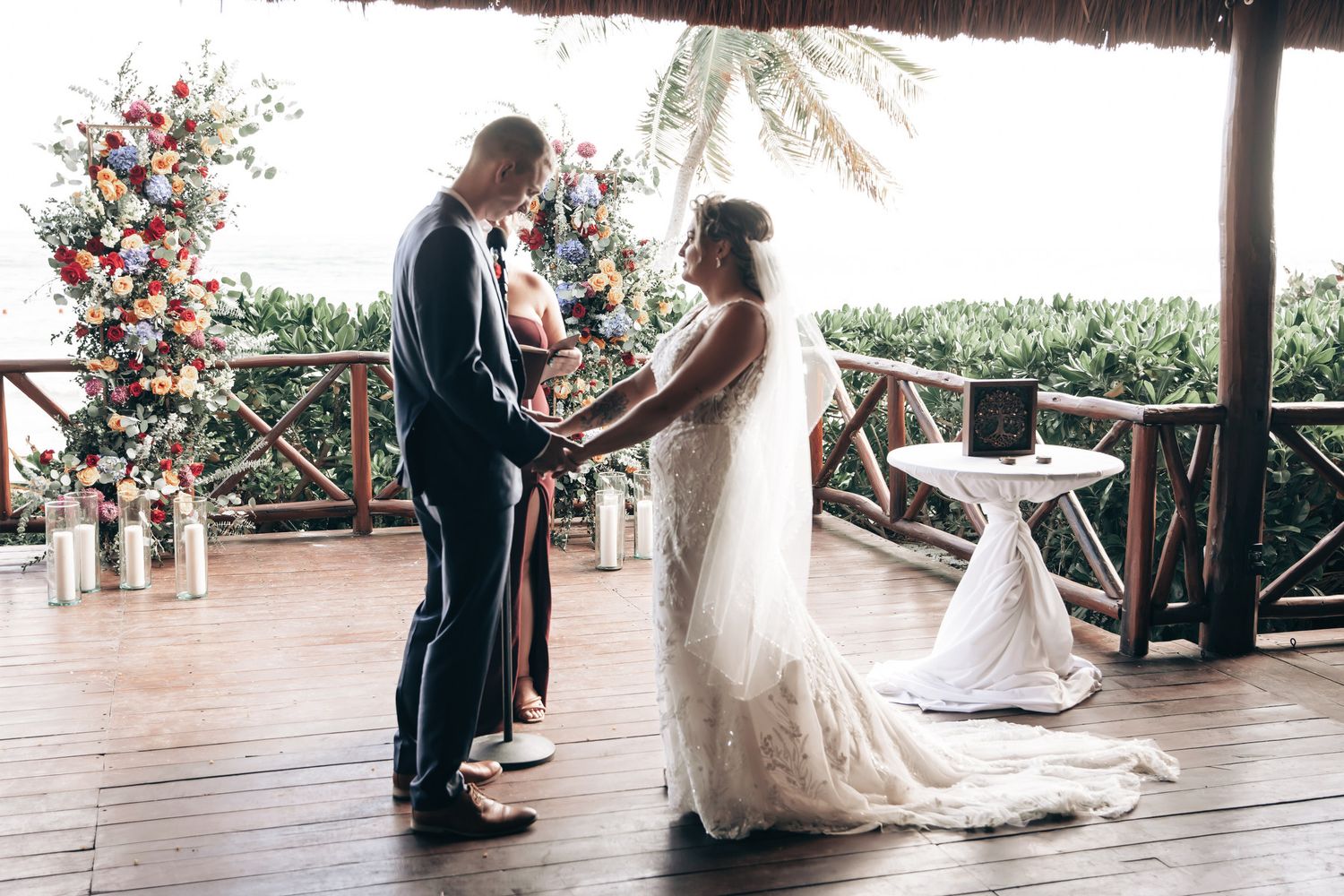 Bride and groom exchanging vows at a tropical outdoor wedding ceremony with a colorful floral arch backdrop.