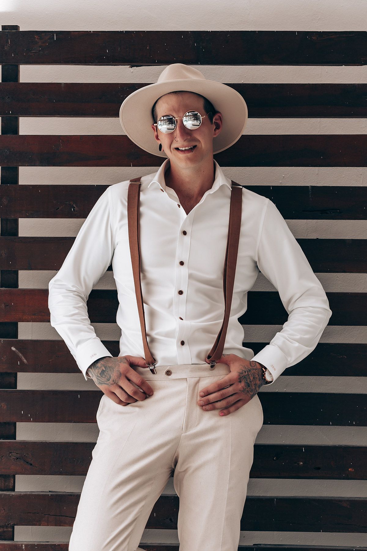 A person in a stylish white outfit with brown suspenders poses against a striped wall background.