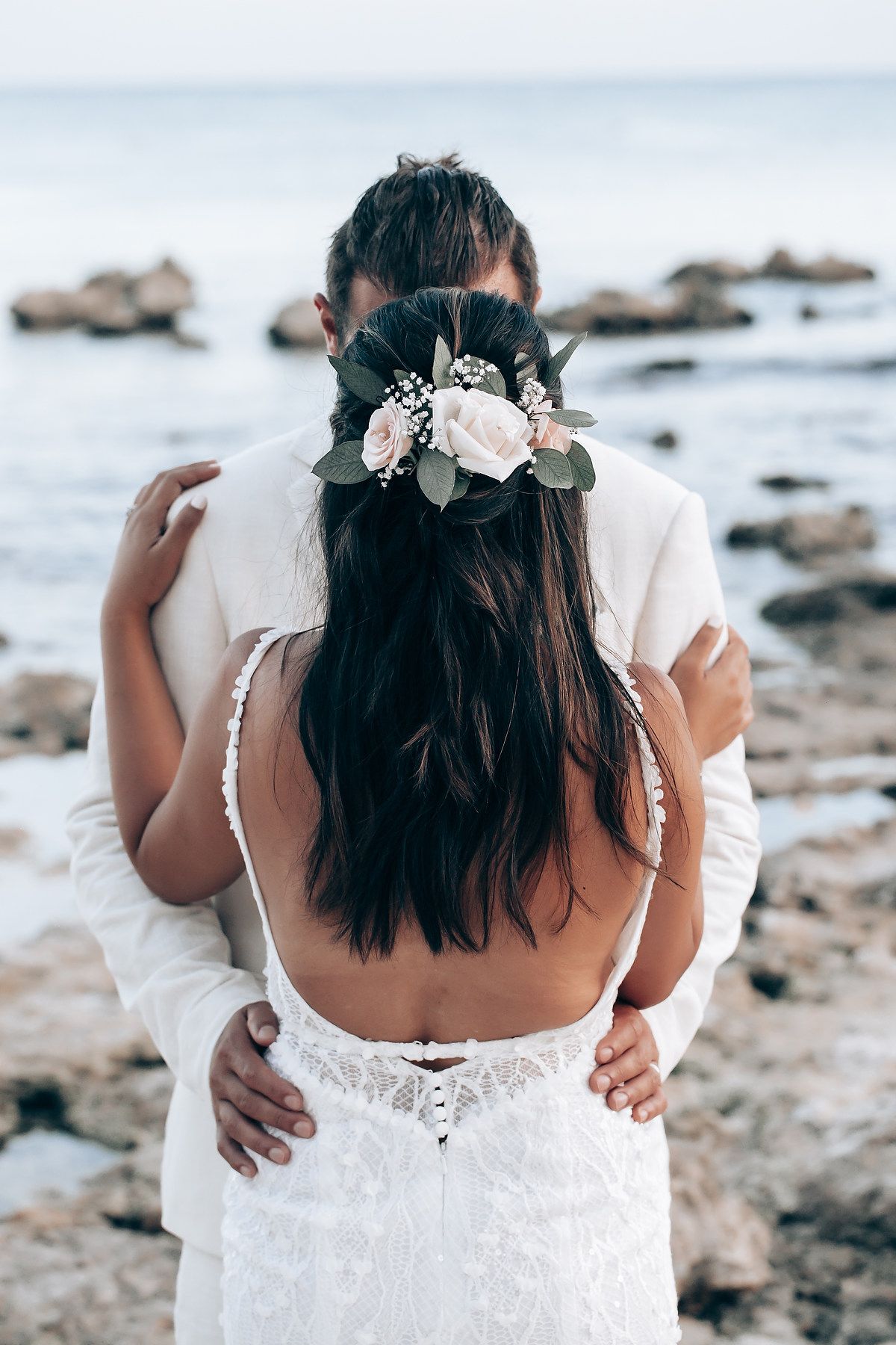 Close-up of bride wearing floral crown and white lace dress with open back design on rocky beach.