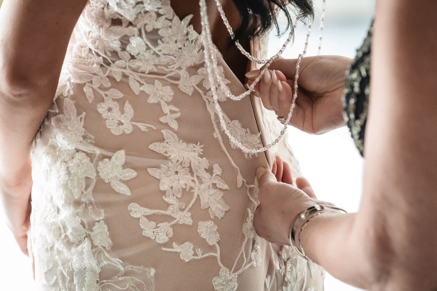 Close-up of bride being buttoned into a floral lace wedding gown with pearl details.