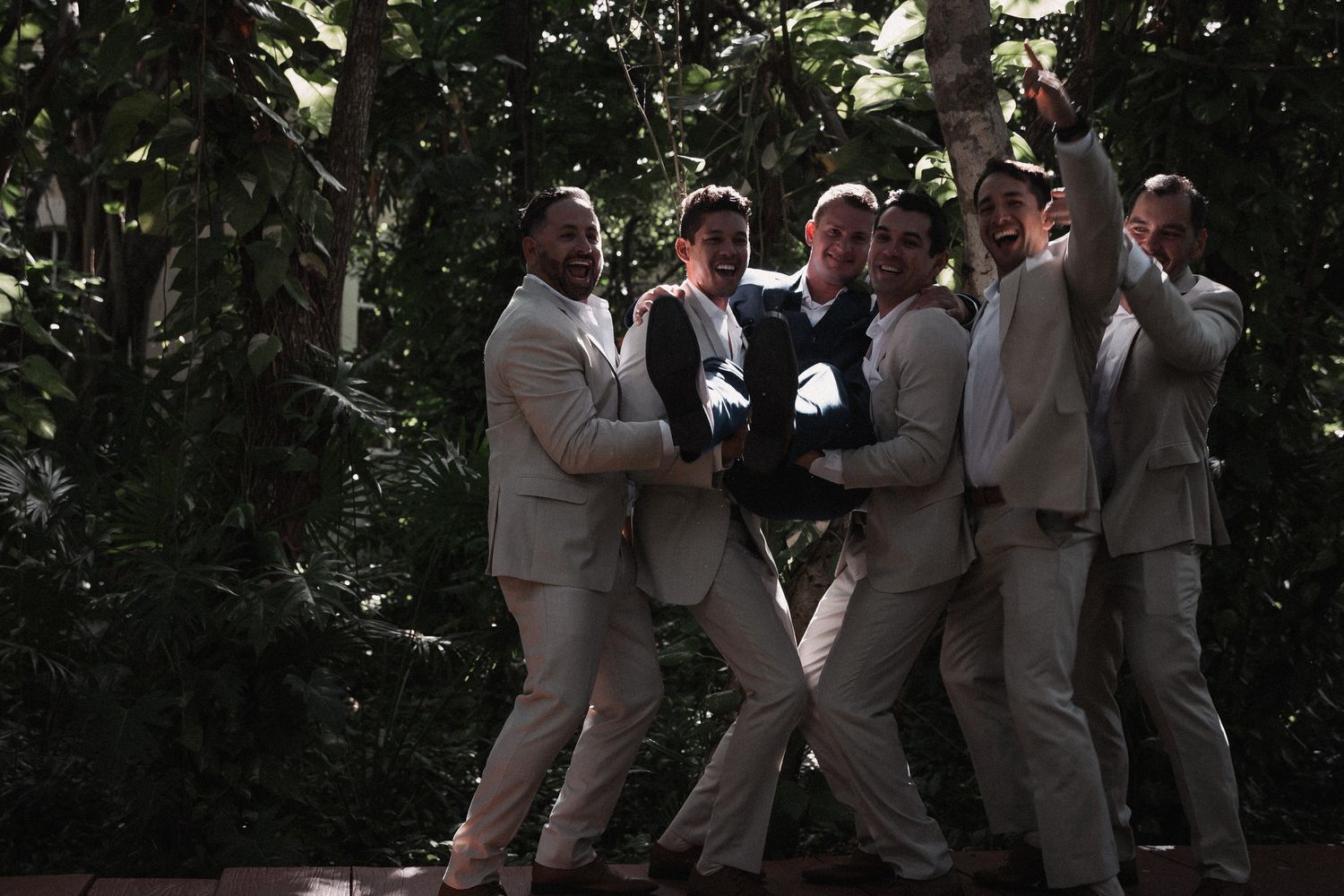 Groomsmen in tan suits playfully lifting groom in lush tropical garden before wedding ceremony.