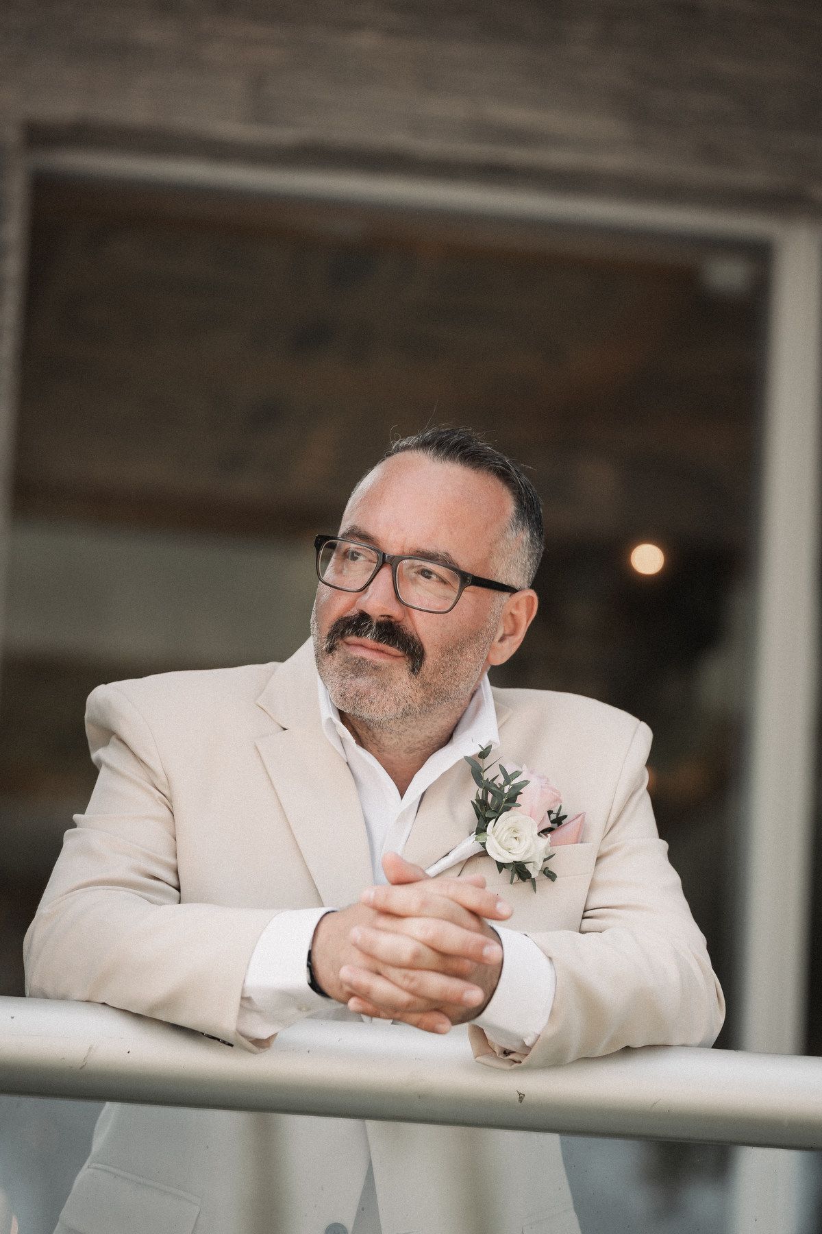 Groom in beige suit with floral boutonniere leaning on railing with thoughtful expression.