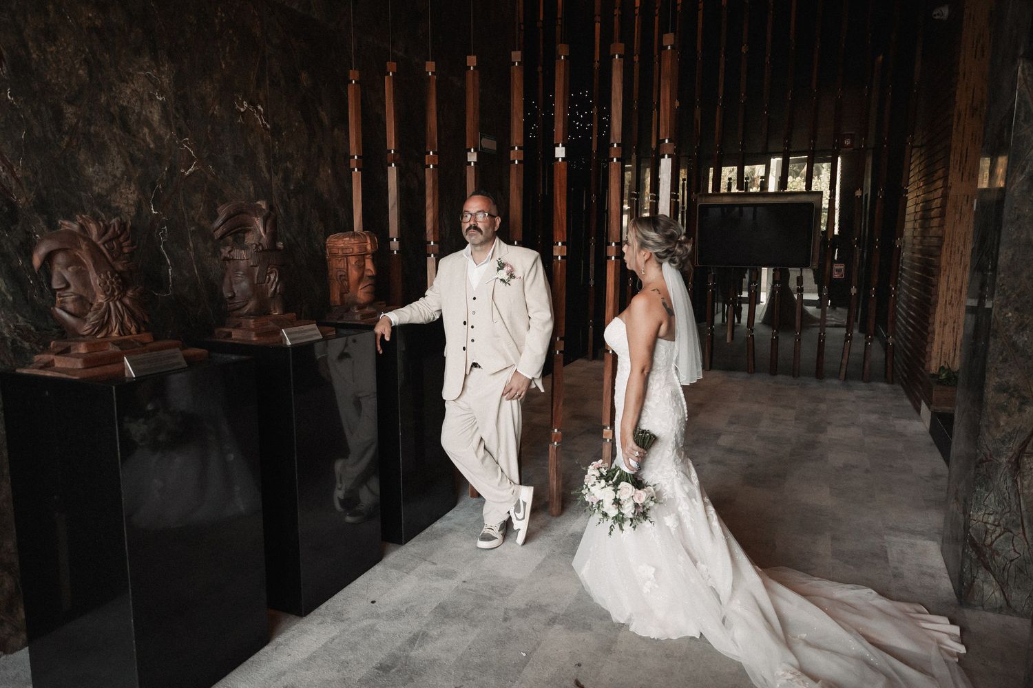 Bride and groom share a joyful moment inside an elegant venue with wooden sculptures and dim lighting.