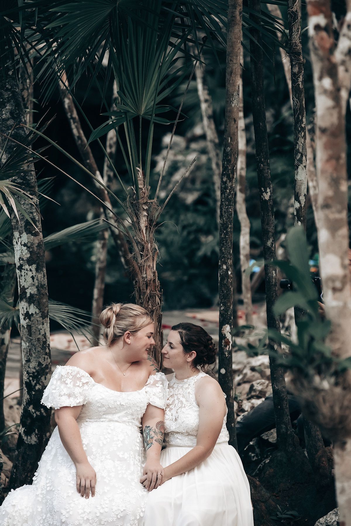 Two brides in white wedding dresses embracing each other in a tropical forest setting.