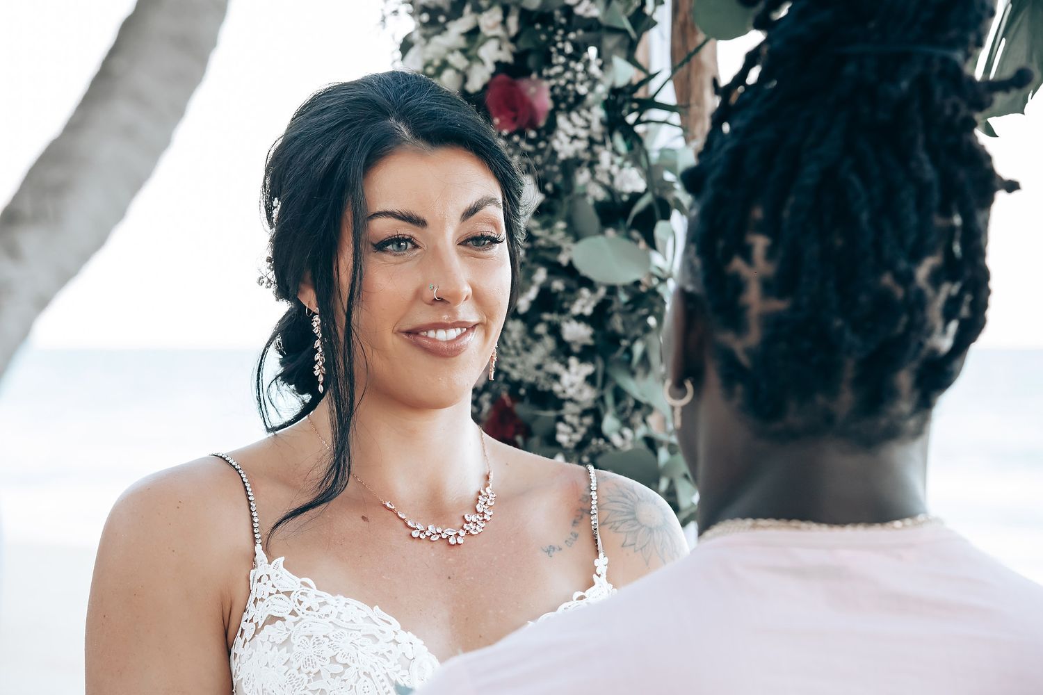 The image shows a smiling bride in a white wedding dress standing with a groom on a tropical beach.