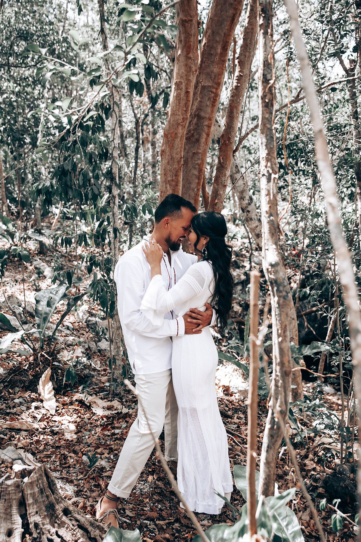 A romantic embrace between two people in white clothing among tall forest trees.