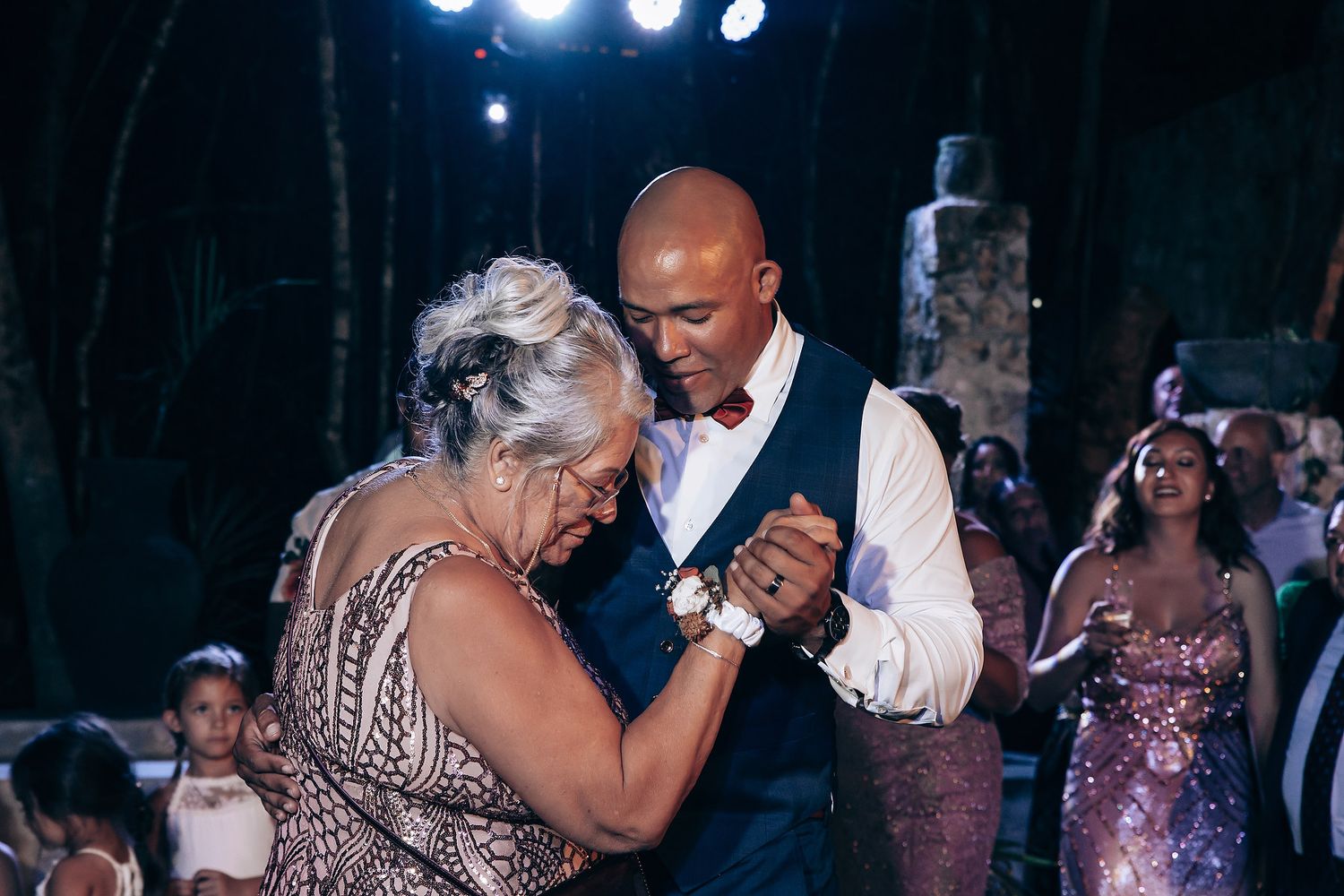 A couple dances together at an evening celebration with sparkling lights and festive decorations in the background.