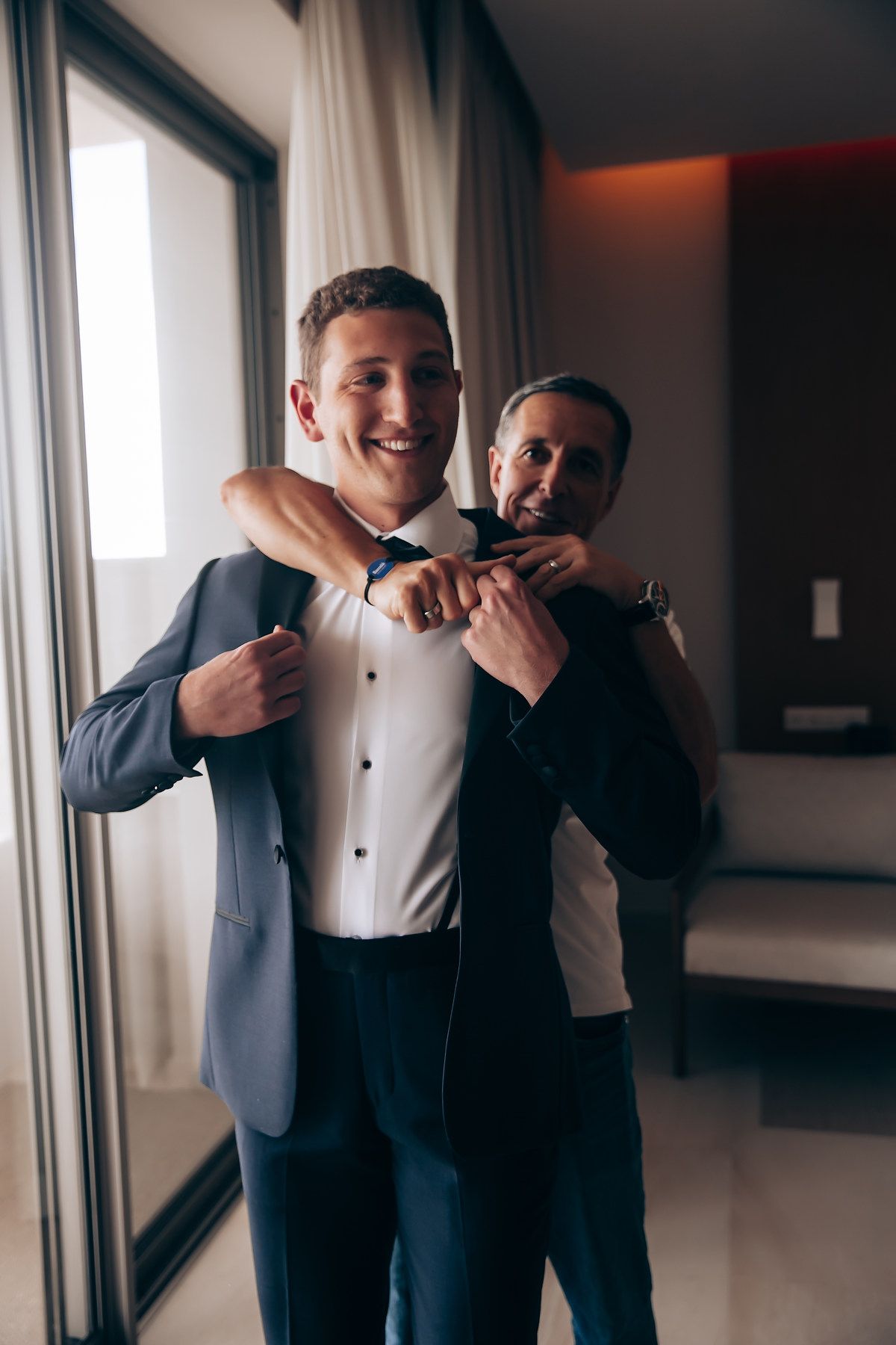 Two people in formal attire share a candid laughing moment together near a window.