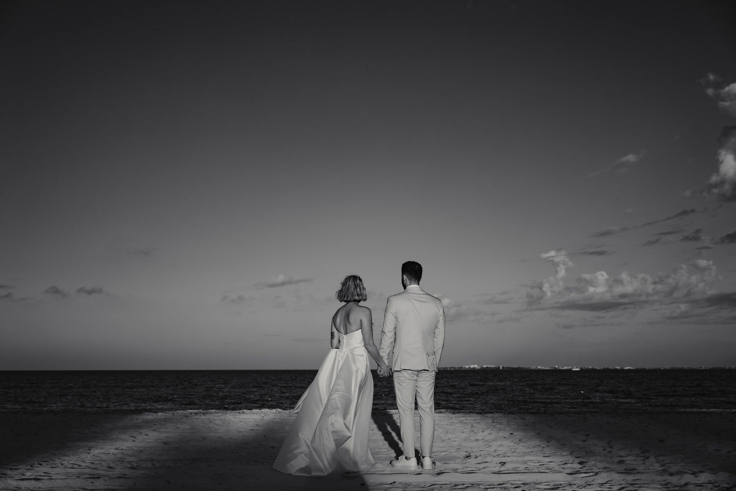 Bride and groom holding hands walking on a beach at sunset in a romantic black and white wedding photo.