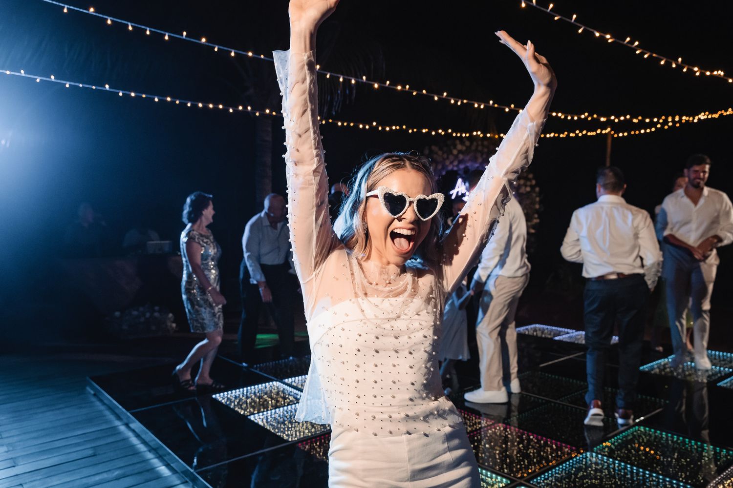 Bride dancing joyfully in sunglasses on a lit dance floor at an outdoor evening wedding reception.
