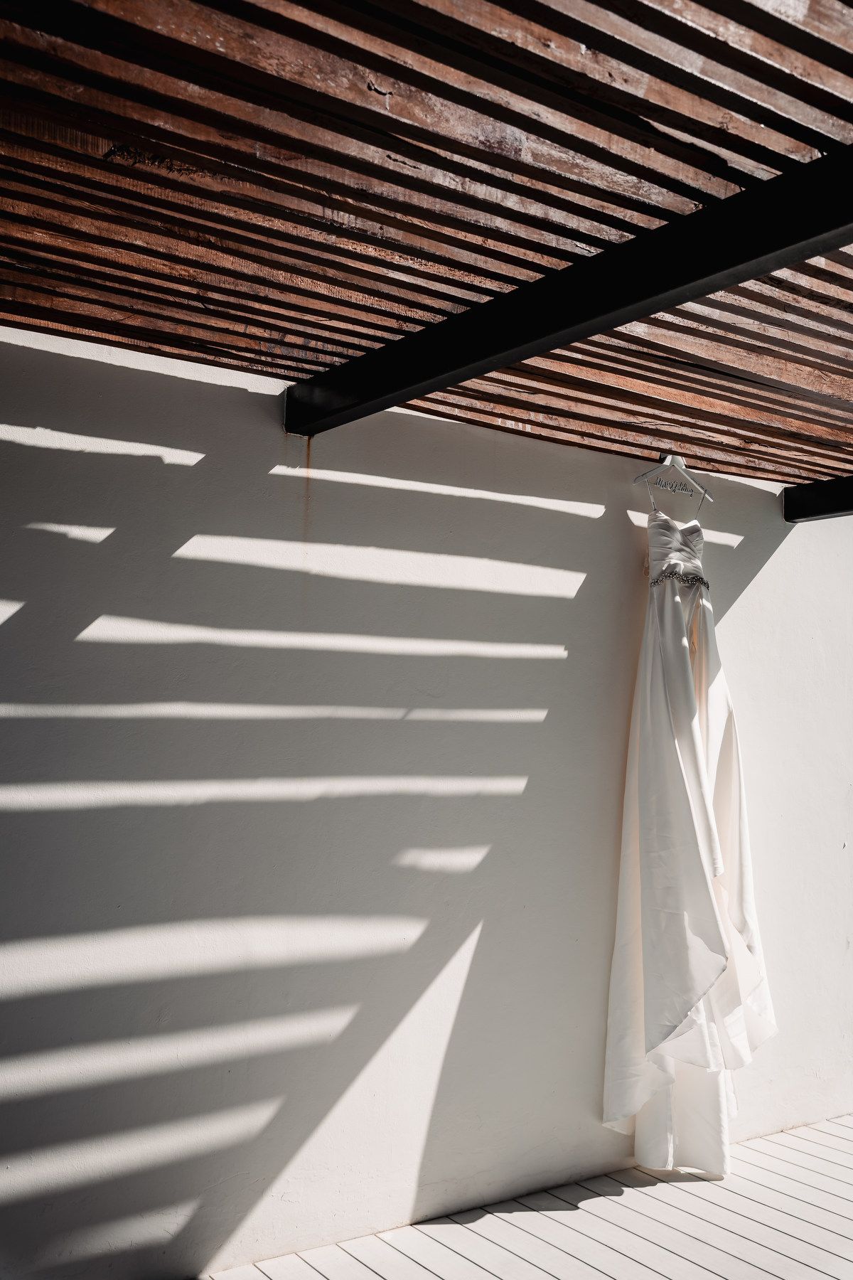 White wedding dress hanging on a sunlit white wall with dramatic wooden slat shadows.