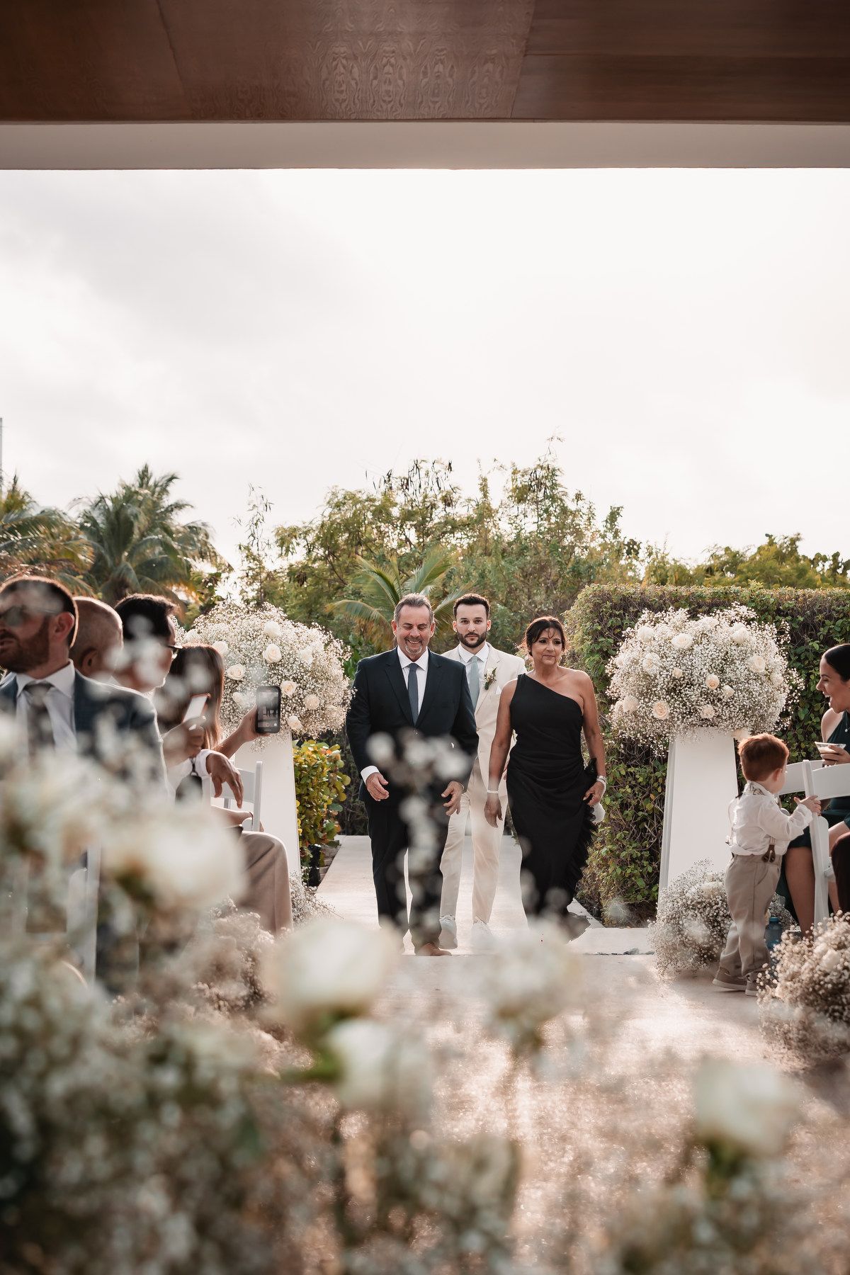 Couple walking down the outdoor wedding aisle lined with white floral arrangements and guests.