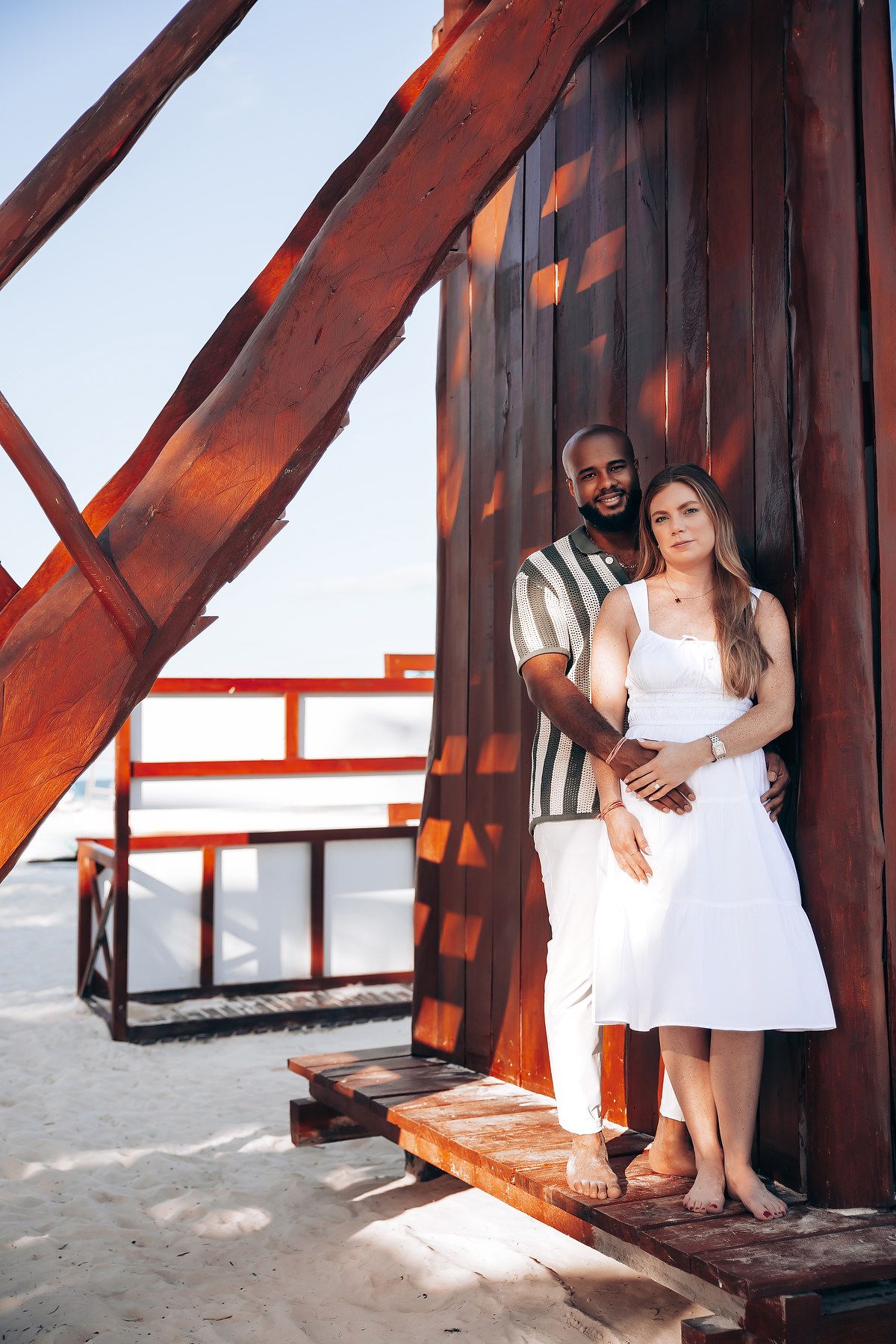 Interracial couple posing together against a rustic wooden beach structure with bright blue sky behind.
