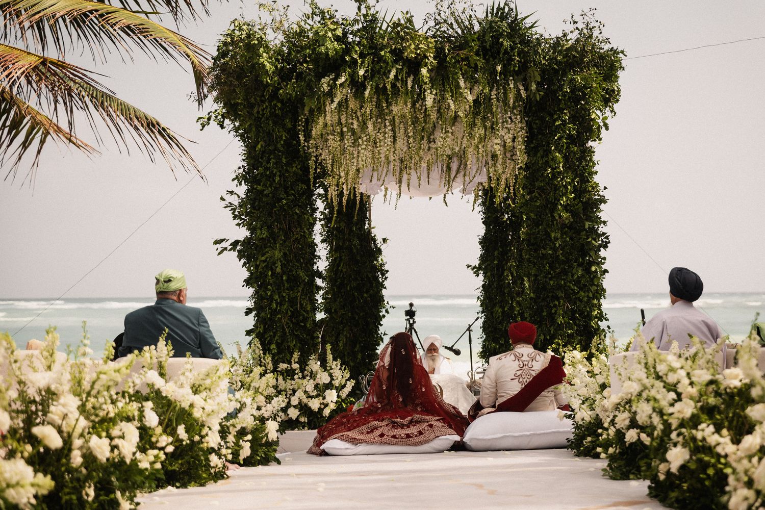 Couple seated under green leafy arch at beach wedding ceremony surrounded by white floral arrangements.