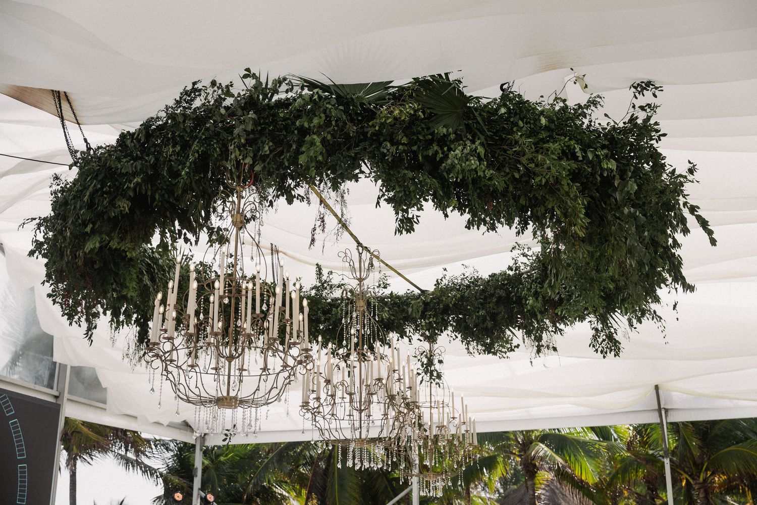 Ornate crystal chandelier surrounded by lush circular greenery wreath hanging from white tent ceiling at wedding reception.