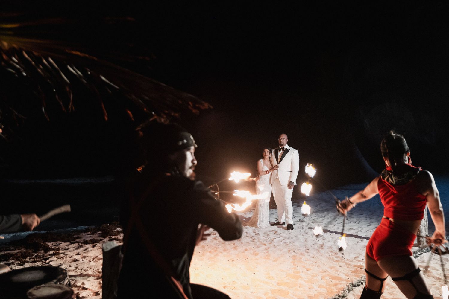 Wedding couple in white posing on beach at night surrounded by fire performers and torches.