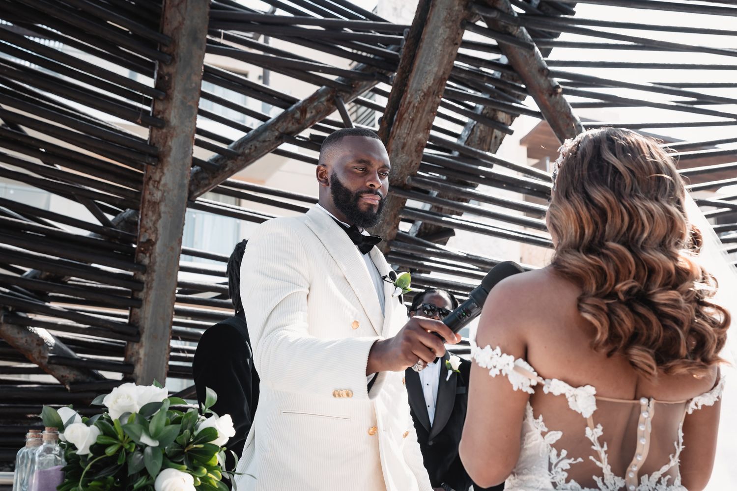 Groom in white tuxedo places ring on bride's finger during outdoor wedding ceremony.
