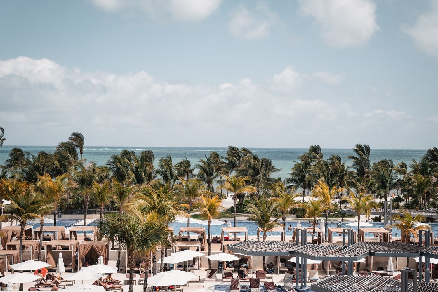 Aerial view of a tropical beach resort with palm trees, pools, and ocean views under a partly cloudy sky.