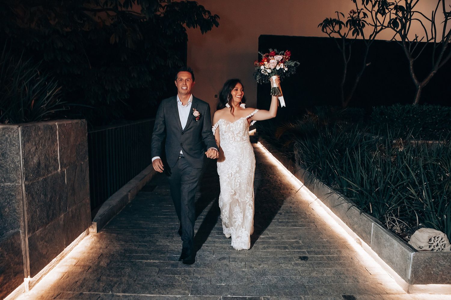 Wedding couple walks along illuminated pathway at night with string lights creating romantic ambiance.