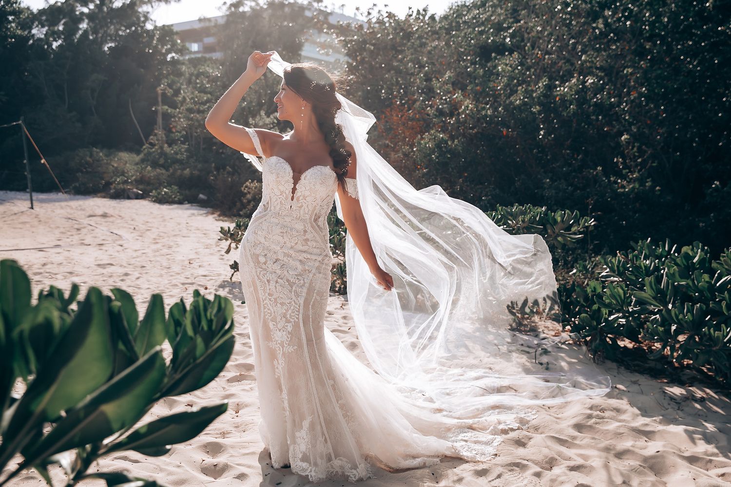 Flowing white wedding dress trails elegantly across a sandy beach with tropical foliage in the background.