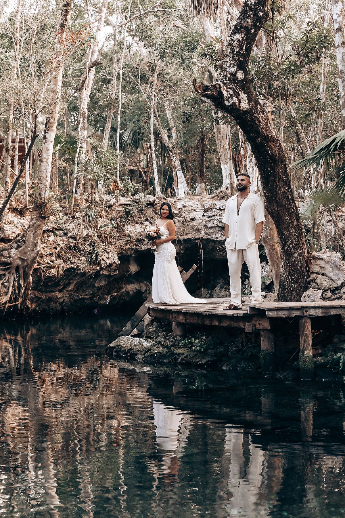 A serene waterside photoshoot with white-clad figures posed along a natural rocky shoreline.