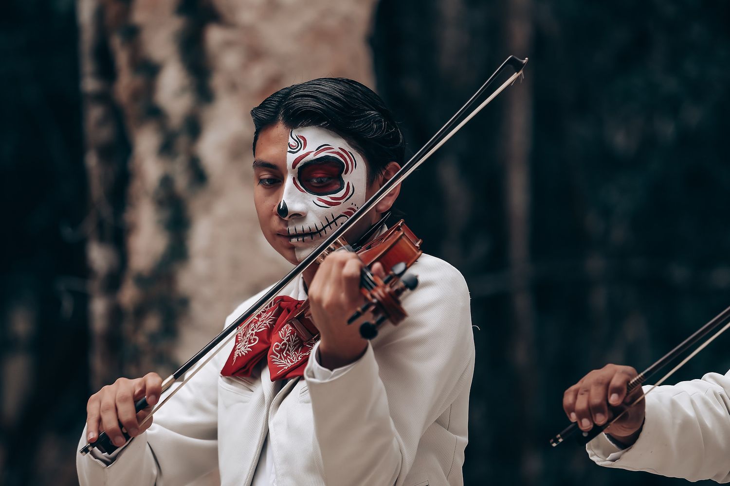 Musician in white suit and red bowtie plays violin in dramatic lighting against dark forest backdrop.
