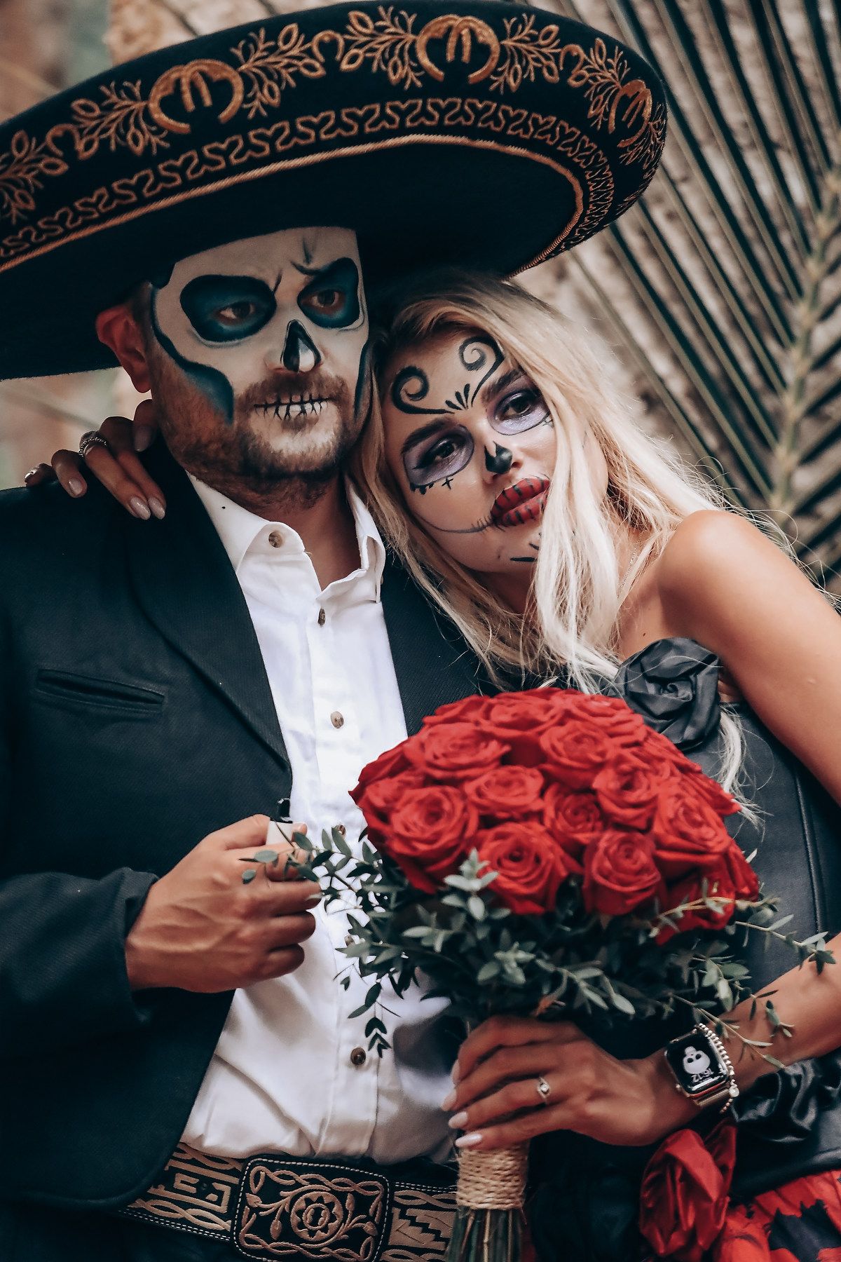 A couple in Day of the Dead makeup poses intimately with a bouquet of red roses against a dark background.