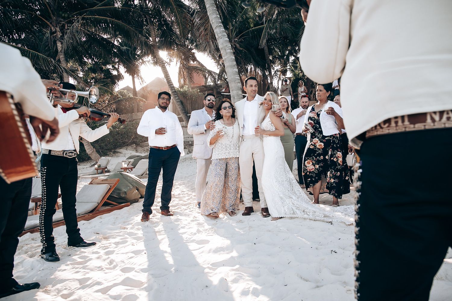 Beach wedding ceremony with guests gathered on white sand path between palm trees.