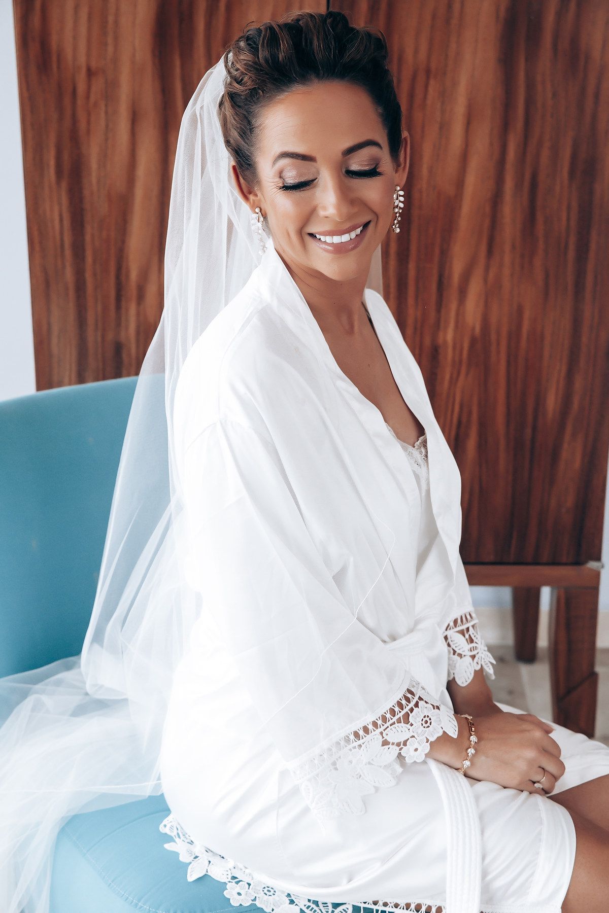 A bride in a white robe smiles joyfully while getting ready on her wedding day.