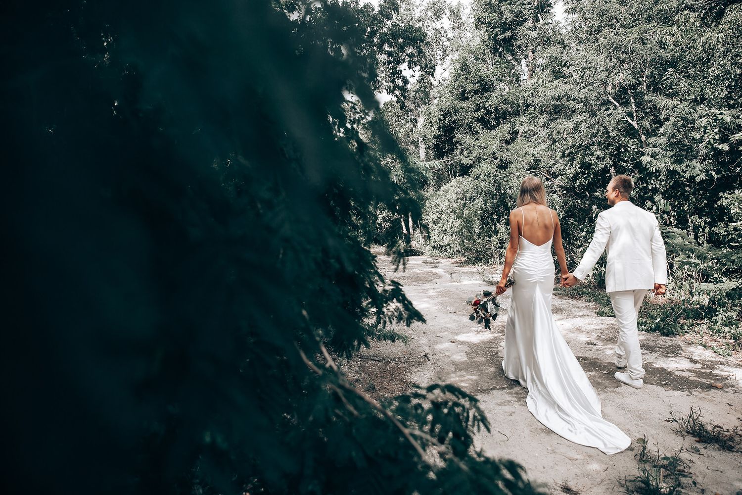 A couple in wedding attire walking down a sunlit garden path holding hands.