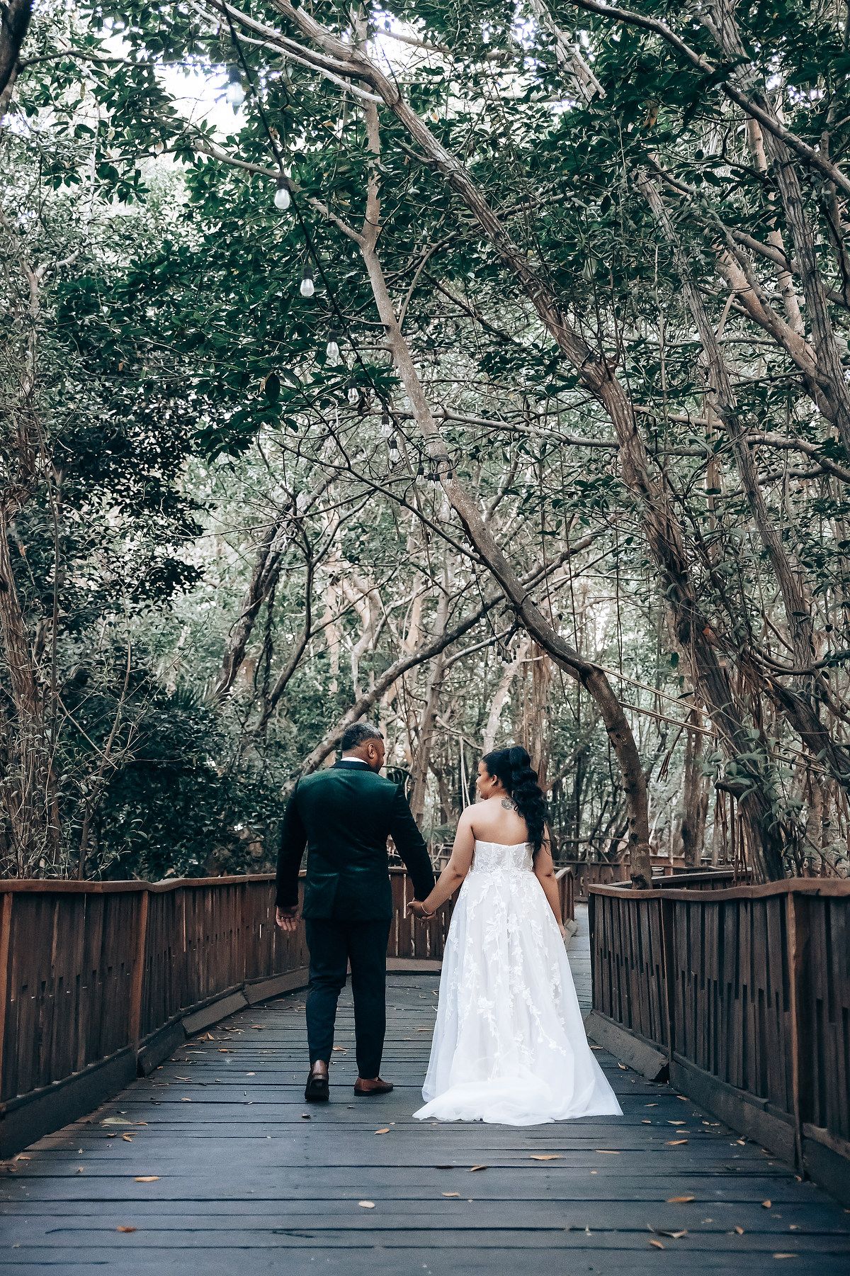 A couple walks along a wooden boardwalk surrounded by lush tropical trees.