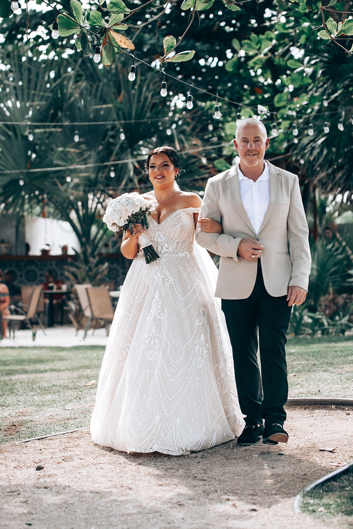 A wedding couple walks together on a sandy path surrounded by tropical greenery.