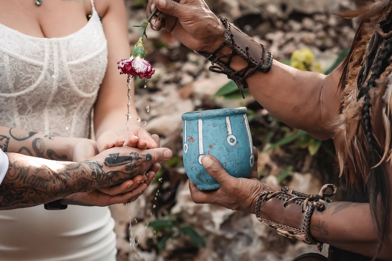 Tattooed hands hold a flower over a turquoise cup in a bohemian outdoor ritual ceremony.