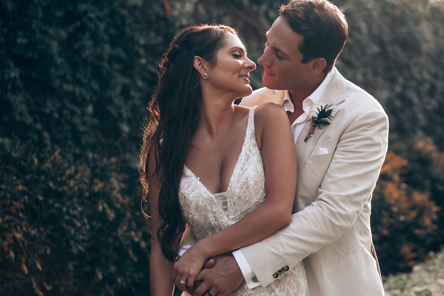 Romantic newlyweds embrace outdoors in elegant wedding attire against a natural backdrop.