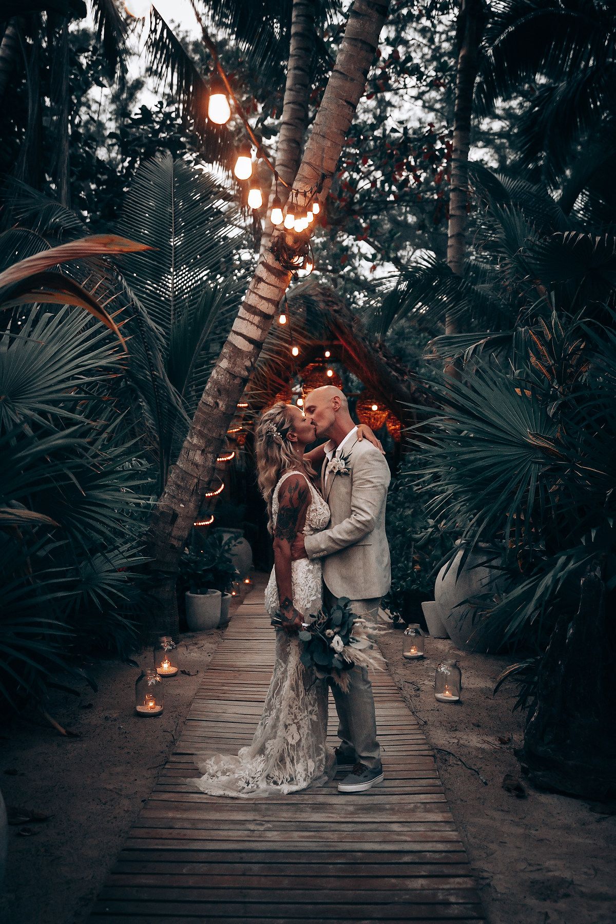 A romantic evening moment on a wooden walkway lit by string lights among tropical palms.