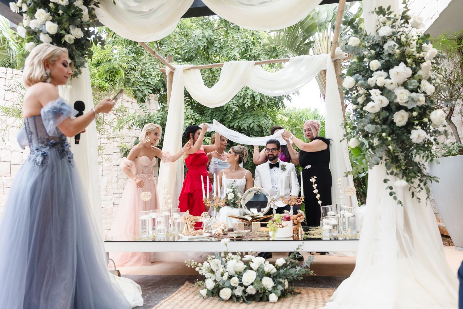 Elegant outdoor wedding ceremony under floral arch with bride, groom, and guests in formal attire.